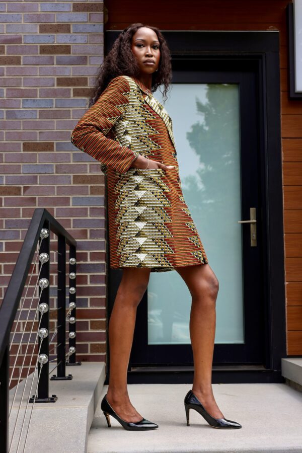 A woman in a brown and gold dress standing next to a brick wall.