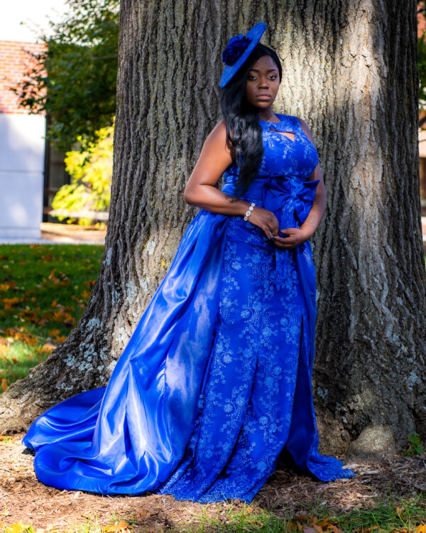 Woman in blue dress and hat by tree.