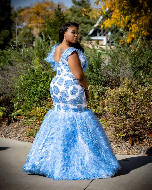 A woman in blue dress standing on sidewalk next to bushes.